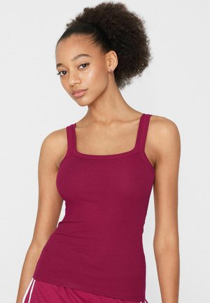 Junge Frau mit lockigem Haar in einem hohen Dutt, die ein eng anliegendes ärmelloses bordeauxrotes Tanktop und bordeauxrote Hosen mit weißen Streifen trägt.