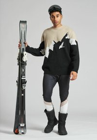 Gestickter Skipullover in Schwarz und Beige mit Bergdesign, kombiniert mit schwarzen Skihosen und isolierten schwarzen Winterstiefeln, während er Skier hält.