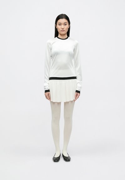 Haut blanc à manches longues avec des accents noirs, associé à une jupe plissée de couleur crème. Le mannequin porte des collants crème et des chaussures plates noires.