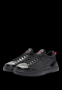 Baskets en cuir noir avec des accents texturés, dotées de lacets noirs, d'un bout arrondi lisse, et d'un détail logo rouge sur la languette et le talon.