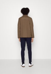 Brun quiltad jacka med horisontella linjer, ståkrage och bakfickdetalj. Matchas med marinblåbyxor och vita skor.