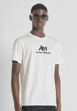 Hombre con camiseta blanca con el logo "ANTONY MORATO" y un diseño geométrico, llevando gafas de sol oscuras, de pie frente a un fondo blanco.