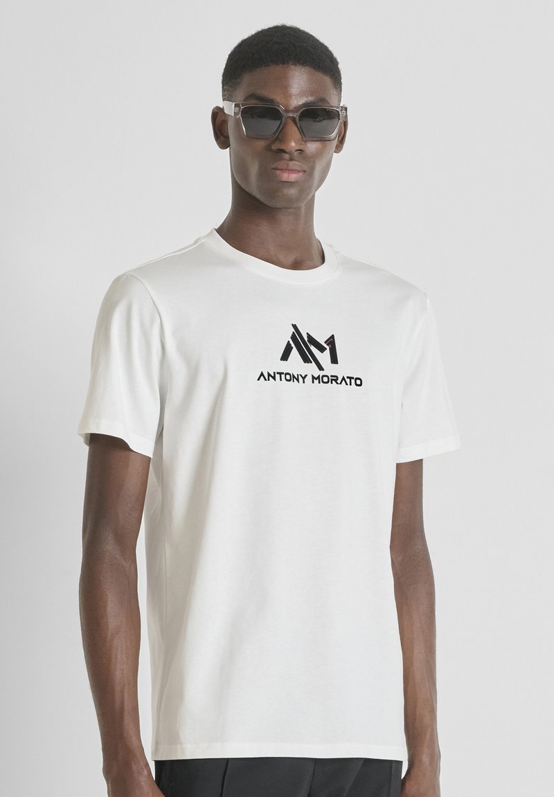 Muškarac u bijelom majici s logotipom "ANTONY MORATO" i geometrijskim uzorkom, u kombinaciji s tamnim sunčanim naočalama, stoji naslonjen na bijelu pozadinu.