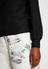 Top nero con motivo esagonale testurizzato, polsini aderenti, abbinato a jeans chiari con effetto scolorito e tie-dye.