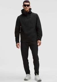 Sort pullover med høj krave og frontlommer med lynlås, parret med sorte bukser og hvide sneakers. Glat materiale med struktureret design.