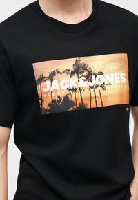 T-shirt noir avec un graphisme de coucher de soleil et de palmier, texte "JACK & JONES DEPUIS MILLE NEUF CENT QUATRE-VINGT-DIX" à l'avant.
