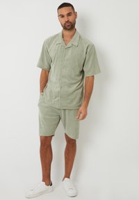 Chemise à manches courtes vert clair à boutons et short assorti en tissu doux. Porté avec des chaussures de sport blanches. Design simple.