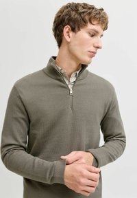 Pull gris-vert à fermeture éclair quart, en tissu texturé, col côtelé, poignets droits, superposé sur une chemise rayée claire.