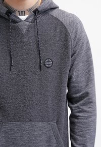 Hoodie gris texturé avec des cordons noirs et un logo circulaire Volcom sur la poitrine, porté par une personne avec un tatouage de chiffres romains sur le cou.