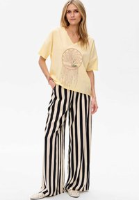 Geel V-hals T-shirt met een cirkelvormige boomafbeelding, gecombineerd met zwart en crèmekleurige verticaal gestreepte wijde broek. Textuurstof, ontspannen pasvorm.