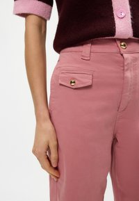 Ροζ βαμβακερό παντελόνι χαρακτηριστικό με μία πλευρική τσέπη και χρυσό κουμπί. Το υλικό φαίνεται μαλακό με λεία υφή, αναδεικνύοντας μια κομψή εφαρμογή.