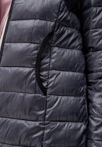 Padded bunda v tmavě námořnické barvě s prošívanou texturou, která má černou podšívku kapes a viditelný zip. Měkký, lesklý materiál.