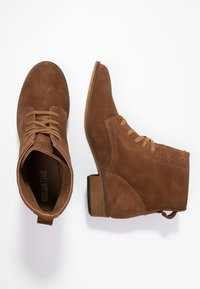Bottines en daim marron avec un bout pointu, un laçage et un talon empilé. Possède une languette à l'arrière et des détails de couture subtils.