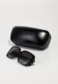 Lunettes de soleil noires carrées avec des verres teintés, étui rigide noir texturé arborant le logo "COACH" en lettres en relief, design minimaliste.