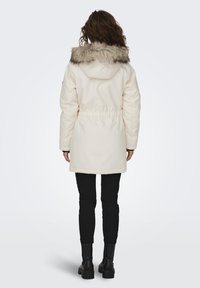 Lichtbeige parka met een nepbont capuchon, een taille met trekkoord en lange mouwen, gecombineerd met zwarte slim-fit broek en enkellaarsjes.