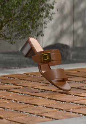 Ruskea korollinen sandaali, jossa kaksi leveää remmiä, kultainen koristeellinen solki, leijuu teksturoidun puisesta päällysteestä ulkona.