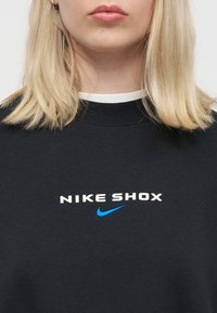 Ξανθός άνθρωπος που φοράει μαύρο φούτερ με λευκό και μπλε λογότυπο "NIKE SHOX" στο κέντρο του στήθους, με μονόχρωμο φόντο.