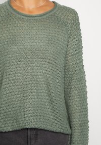 Vihreä neulepaita, jossa on avoin kudontamallia, pyöreä kaula-aukko ja pitkät hihat, yhdistetty tummiin housuihin. Työvaiheet korostavat ilmavaa rakennetta.