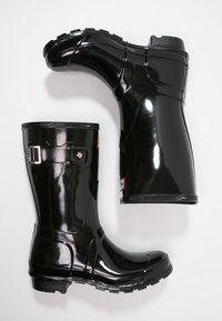 Bottes de pluie en caoutchouc noir brillant avec un bout arrondi, une semelle texturée, une boucle sur le côté et une étiquette de logo près de la cheville.