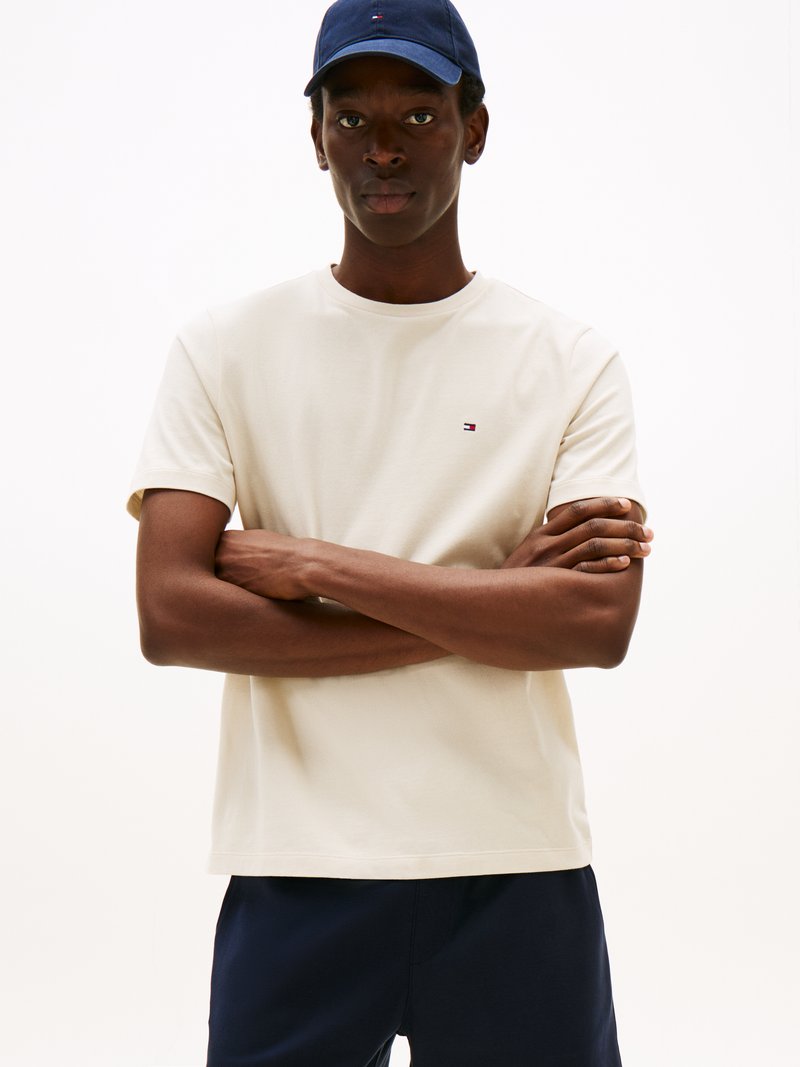 Beige short-sleeve T-shirt with crew neckline, featuring a small logo on the chest. Worn with navy pants and a matching cap.