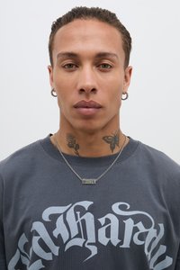 T-shirt grigia con testo bianco audace, collana di catene d'argento sovrapposte con un pendente di metallo e tatuaggi visibili sul collo.