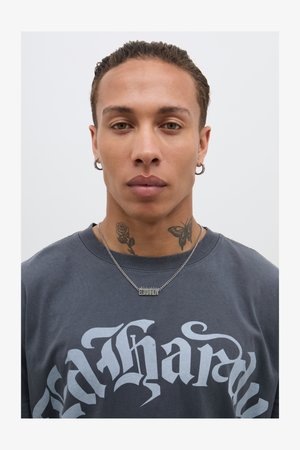 Szürke póló merész fehér szöveggel, többrétegű ezüst lánc nyaklánc fém medállal, és látható tetoválások a nyakon.