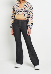 Pull-over à motif flou en crème, rose, bleu et feuillé avec une demi-fermeture éclair. Associé à un jean noir évasé taille haute et des baskets blanches.