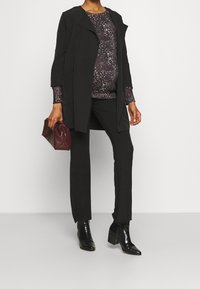 Zwangere persoon in zwarte broek, zwarte jas, bedrukt donker top, zwarte haklaarzen, met een kleine bordeauxrode handtas.