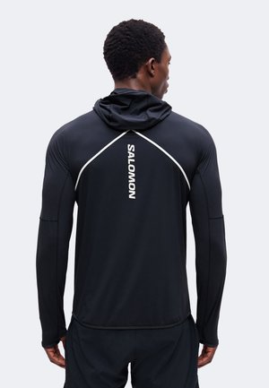Mann trägt schwarzen langärmligen Sport-Hoodie mit "SALOMON" Logo am oberen Rücken und lichtreflektierendem Besatz über den Schultern.
