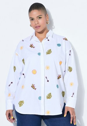 Osoba s krátkými vlasy, která má na sobě bílou košili s barevnými vyšívanými vzory listů, slunce, ryby a citrusů, doplněnou modrými džínami.