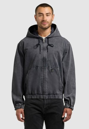 Vyras dėvi tamsiai pilką, su užtrauktuku užsegamą gobtuvinį džemperį su priekinių kišenių ir elastinių rankogalių detalėmis, derina jį su juodomis kelnėmis, stovi priešais vienspalvį foną.