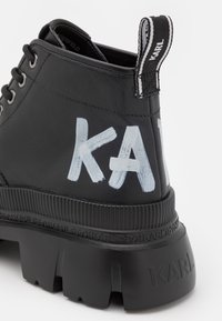 Botín negro de cuero hasta el tobillo con suela gruesa, con la marca "KARL" en el talón y en la lengüeta, presentando grandes letras blancas pintadas en el lado.
