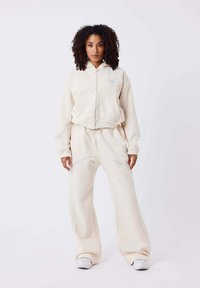 Beige Fleece-Jacke mit Reißverschluss und passenden weit geschnittenen Hosen, mit hellblauen Akzentnähten und einem Logo auf der Brust.