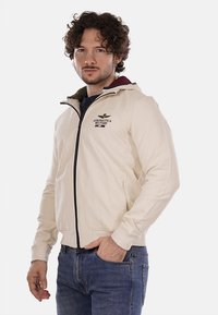 Giacca zip-up color crema in un tessuto leggero, con cappuccio, dettagli del logo ricamato e zip nera. Indossata con jeans blu.