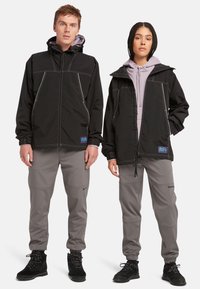 Chaquetas exteriores negras con costuras blancas en contraste, combinadas con pantalones gris claro. Las chaquetas cuentan con capuchas y acentos de marca.