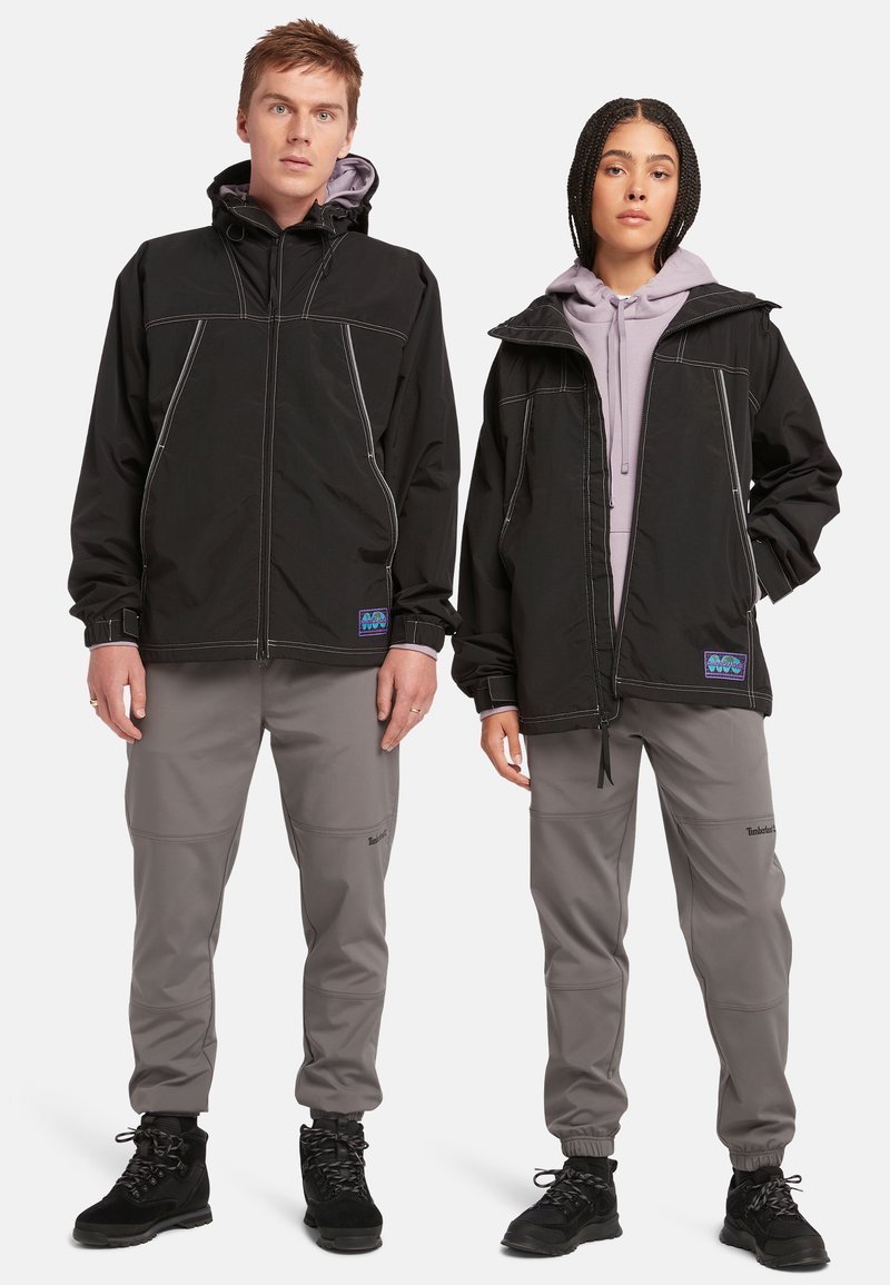 Chaquetas exteriores negras con costuras blancas en contraste, combinadas con pantalones gris claro. Las chaquetas cuentan con capuchas y acentos de marca.