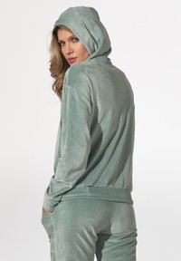 Jasnozielona bluza z weluru z kapturem i długimi rękawami. Miękka faktura i luźny krój. Widok z tyłu, bez widocznych zapięć czy wzorów.