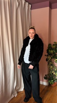 Νέα γυναίκα με ξανθό κότσο, φορώντας μαύρο γούνινο μπουφάν, λευκό τοπ, μαύρο φαρδύ παντελόνι, στέκεται δίπλα σε μπεζ κουρτίνα και γλαστρόφυτο πάνω σε ξύλινο δάπεδο.