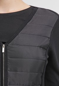 Svart vadderad väst med synlig horisontell sömnad och en silverdragkedja över en svart långärmad skjorta på en person.