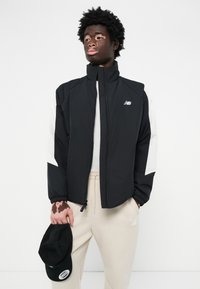 Giacca nera con pannelli bianchi, zip completa, colletto alto e polsini elasticizzati; il modello tiene un cappellino nero e indossa pantaloni beige.
