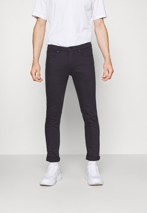 Jeans slim-fit grigio scuro con cinque tasche, cuciture robuste e orlo arrotolato, abbinati a sneaker bianche e una t-shirt bianca semplice.