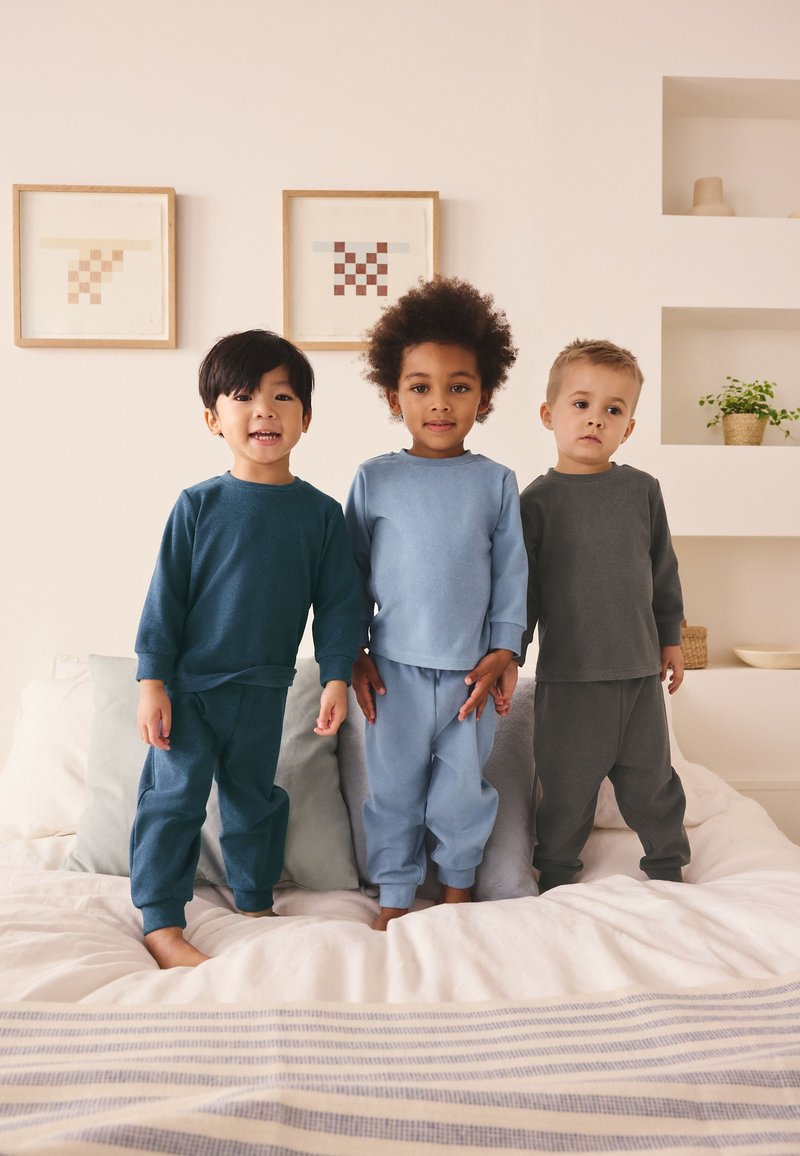 Drei kleine Kinder tragen passende Langarmshirts und Hosen und stehen barfuß auf einem Bett in einem neutral gestalteten Schlafzimmer.
