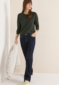Mørkegrøn strikket sweater med lange ærmer, kombineret med mørke denim flare jeans og lyse sneakers. Model holder en lysegrå sweatshirt.
