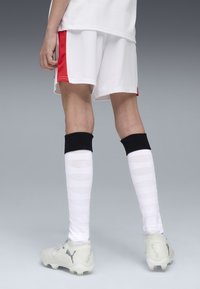 Weiße Fußballshorts mit roten Seitenakzenten und schwarzen knielangen Socken, kombiniert mit weißen Schuhen, die ein strukturiertes Design und kontrastierende Schnürsenkel aufweisen.