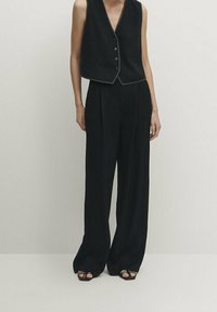 Schwarze ärmellose Weste mit V-Ausschnitt und Knöpfen, kombiniert mit weiten schwarzen Hosen. Glatter Stoff mit elegantem, minimalistischem Design.