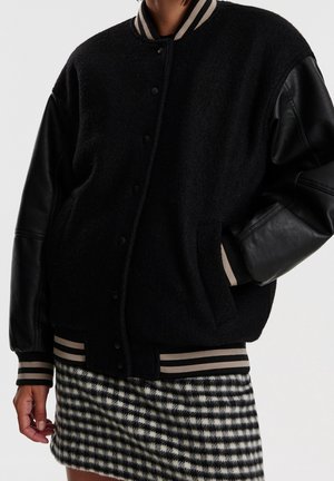 Personne portant une veste varsity noire avec des manches en cuir et des poignets rayés, portée avec une jupe à carreaux noire et blanche sur un fond neutre.
