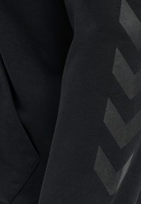 Sweatshirt noir avec une texture douce, une coupe décontractée et des motifs chevrons noirs angulaires le long de la manche droite.