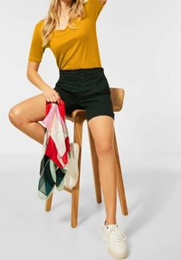 Žlutý top s krátkým rukávem a černé šortky. Model sedí na dřevěném stoličce a drží pestrobarevný šátek. Na nohou má bílé tenisky.