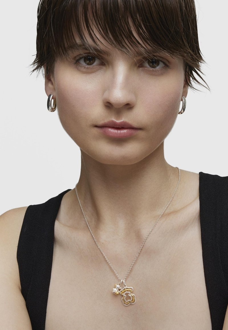 Collana in argento con pendente a forma di fiore dorato e un accento di perla. La modella indossa un top nero e orecchini a cerchio in argento.