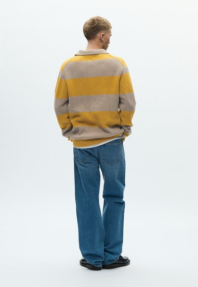 Πλεκτό πουλόβερ με κίτρινες και γκρι οριζόντιες ρίγες, με γιακά, συνδυασμένο με φαρδιά μπλε τζιν και μαύρα παπούτσια.